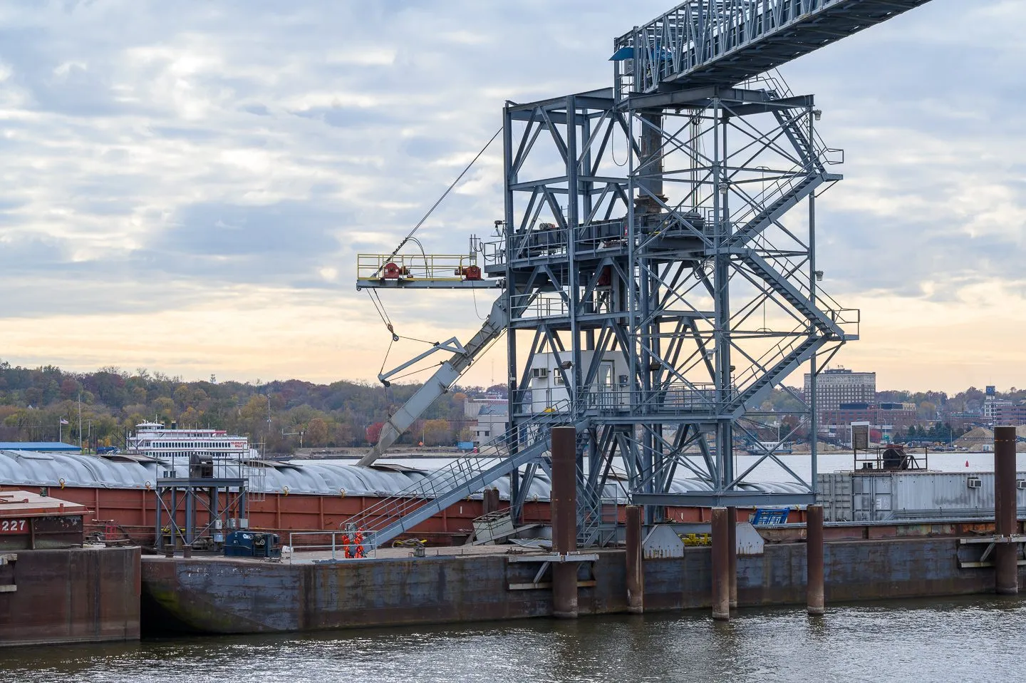 An industrial loading structure transferring material into a barge on a river, with a cityscape and trees in the background.